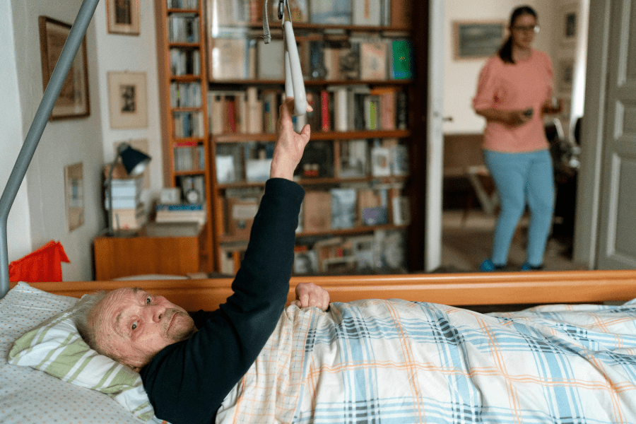 Senior leży w łóżku, a w tle widać idącą do niego opiekunkę.
